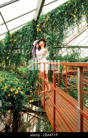 Ein einsam schönes Mädchen mit mehrfarbigen Dreadlocks in einem weißen Kleid steht auf einer roten Treppe in einem Bogen mit Blumen. Gewächshaus mit exotischem tropischem p Stockfoto