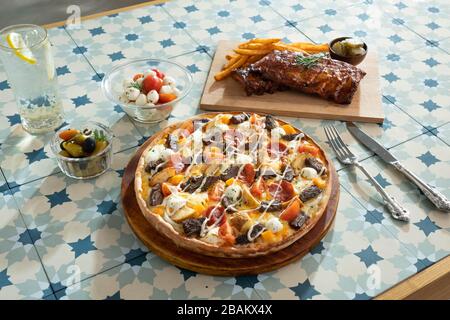 Pfanne Pizza mit Fleisch, Kartoffel, Gemüsebesatz und Schweinerippe und Beilage auf einem Tisch Stockfoto