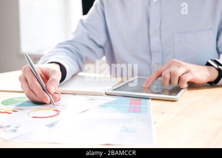Nahaufnahme des nicht erkennbaren Finanziers, der am Schreibtisch sitzt und Tablet verwendet, während er Diagramme im Büro analysiert Stockfoto