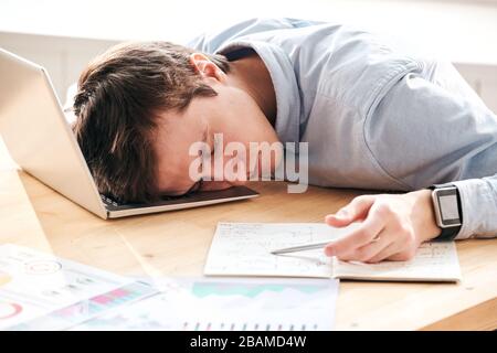 Junger, müder Mann erschöpft mit Berechnungen, die auf dem Laptop liegen, während er am Arbeitsplatz schlief Stockfoto