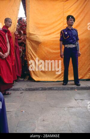Ein Monch-Tänzer erwartet seine Wende, um den Tanzbereich zu betreten, während ein Polizist Wache bei Thimphu, Bhutans jährlichem Tsehu-Festival, steht. (10-10-89) BX50 Stockfoto
