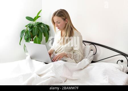 Junge hübsche, blonde Frau aus europa, die von zu Hause aus im Bett mit Laptop und Smartphone arbeitet und studiert, sie ist in leichter, komfortabler Hauskleidung. Stockfoto