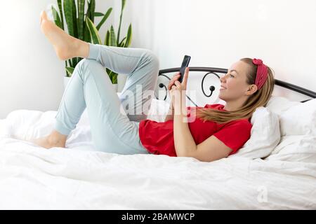 Junge lächelnde Frau mit blonden Haaren in einem roten T-Shirt und bequemen Hosen liegt auf einem weißen Bett mit Smartphone, sie arbeitet, studiert von zu Hause, schreibt A. Stockfoto