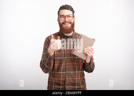 Fröhlicher junger Mann hält ein Notizbuch oder ein Buch, lächelt an der Kamera, zeigt wie Taste oder Daumen auf weißem Hintergrund. Stockfoto