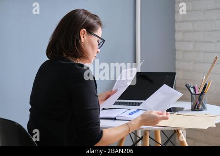 Seriöse Frau, die Geschäftspapiere in Händen hält, Dokumente, die in Briefumschlägen, auf Schreibtischlaptop, Telefon, Terminkalender eingegangen sind Stockfoto