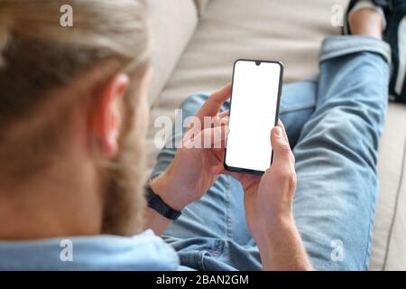 Junger technischer Benutzer, der sich auf dem Sofa entspannt, indem er das Smartphone mit einem leeren weißen Bildschirm verspottete. Stockfoto