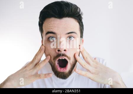 Mann mit schockierter, verblüffter Gesichtsausdrucksweise sieht direkt auf der Kamera aus. Finger mit geöffnetem Mund an Wangen halten. Isoliert über weißem Hintergrund. Stockfoto