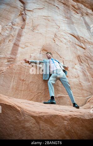 Der nervöse Geschäftsmann klammert sich an ein Felswand und balanciert auf einem schmalen Felsvorsprung in einem roten Felsschlucht Stockfoto