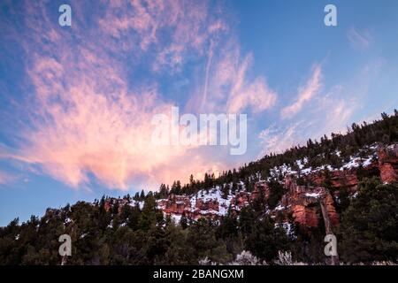 In einem abgelegenen Wüstenschlucht in Utah sind rote Klippen im Winter schneebedeckt. Überhängende Whiskywolken leuchten bei Sonnenuntergang in Rosa auf. Stockfoto