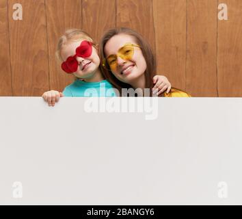 Lächelnde glückliche Familie, die hinter einer weißen Leerplatte vor Holzhintergrund steht. Lustige Gesichter. Hinter einem Banner, leerer Platz für einen tex, wird ein Peeking durchgeführt Stockfoto