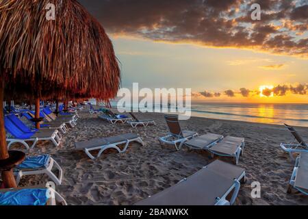 Goldener Sonnenaufgang über Reihen von Lounge-Stühlen und Palmenschirmen an einem sandigen Strandurlaub in der Karibik an der Riviera Maya in Cancun, Mexiko. Stockfoto