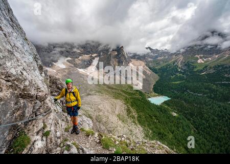 Junger Mann, Bergsteiger auf einer festen Seilstrecke, über Ferrata Vandelli, Blick auf Lago di Sorapis, Sorapiss-Rundkurs, Berge mit niedrigen Wolken, in die Dolden Stockfoto