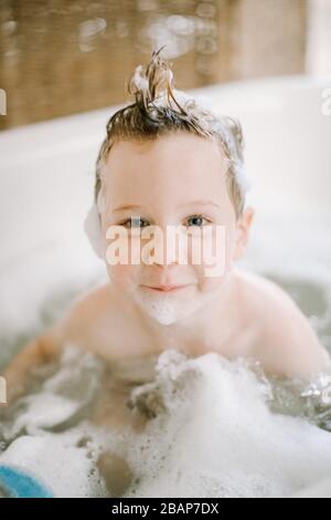 Vorschuljunge in Badewanne mit Blasen im Gesicht und Haaren, die auf die Kamera blicken und lächeln Stockfoto