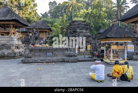 Junge balinesische Mutter, Vater und Tochter in heller traditioneller Kleidung knieend vor dem Tempelaltar und betend zu Vishnu. Stockfoto