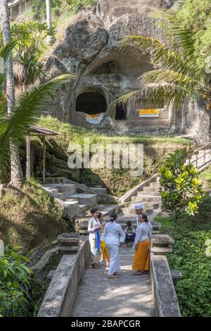 Drei balinesische Hindu-Frauen in traditionellen Sarong-Kleidung stehen und sprechen auf der Tempelbrücke mit Tempelgebäuden und Palmen im Hintergrund. Stockfoto