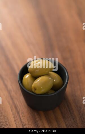 Riesige grüne Oliven in schwarzer Schüssel auf Walnusstisch, flacher Fokus Stockfoto