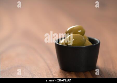 Riesige grüne Oliven in schwarzer Schüssel auf Walnusstisch, flacher Fokus Stockfoto