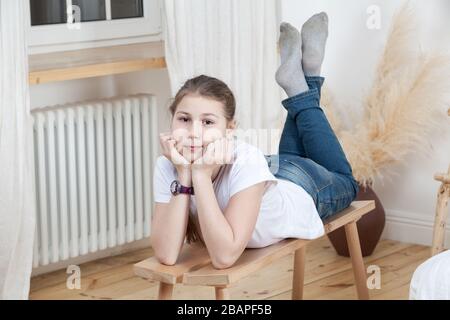 Hübsches kaukasisches Mädchen, das auf Holzbank im Schlafzimmer in der Nähe von Heizung unter Fensterbank liegt, gekleidetes weißes T-Shirt und blaue Jeans Stockfoto