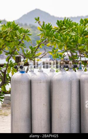 Viele Aluminium-Tauchsauerstofftanks stehen vor den grünen Strandbüschen und dem Meerbusen auf dem Sand in Thailand mit Kopierraum Stockfoto