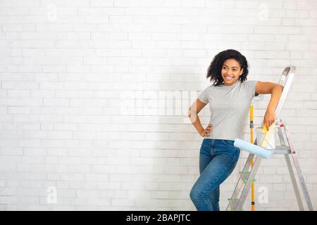 Frau neben Stepladder mit Walze in der Hand auf weißem Backstein-Wandhintergrund Stockfoto