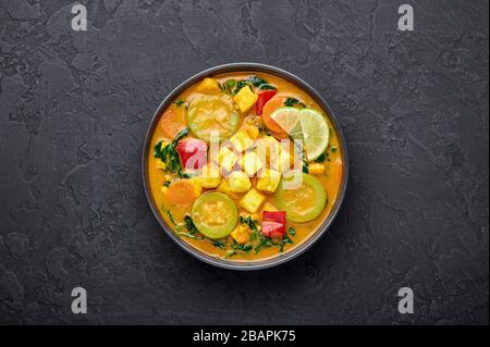 Gelber Veg Thai Curry mit Tofu und Gemüse in schwarzer Schüssel bei dunklem Schieferhintergrund. Vegetarisches Thai-Curry mit Tofu, Zucchini, Pfeffer, Spinat, Auto Stockfoto