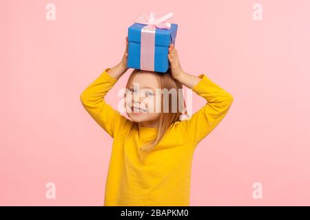 Portrait des fröhlichen kleinen Mädchens, das Geschenkbox auf dem Kopf hält, Spaß mit Geschenk hat und freudig mit der Kamera, dem Geburtstag oder der Weihnachtsfeier lächelt Stockfoto