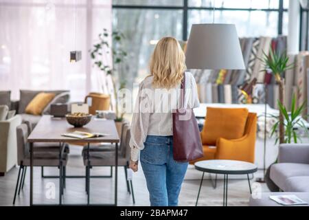 Alles für das Haus. Frau in einer weißen Bluse mit einer burgunderfarbenen Tasche auf der Schulter, die mit dem Rücken im Möbelsalon steht. Stockfoto