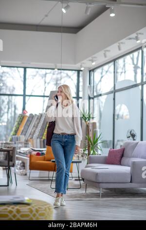 Besuchen Sie den Möbelsalon. Frau mit blonden Haaren, die auf dem Smartphone spricht, zur Seite schaut, im großen Saal spazieren geht, lächelt. Stockfoto