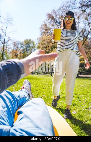 Mann, der auf dem Luftsofa im öffentlichen Park liegt, Frau, die eine Papierbecher mit Tee hält Stockfoto