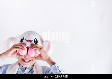 Lächelnder Junge in einer flauschigen Maske aus Osterhase verschließt seine Augen mit handgeschmalten rosafarbenen Eiern vor hellgrauem Hintergrund. Frohe Ostern Karte. Stockfoto