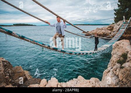 Paar Frau mit Mann zu Fuß durch wackelige Hängebrücke überqueren Sea Bay Stockfoto