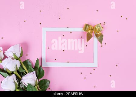 Rosa Rosen Blumen mit goldenem Bogen, weißem Rahmen und goldenen Glitzersternen auf pinkfarbenem Hintergrund, flacher Lay Stockfoto