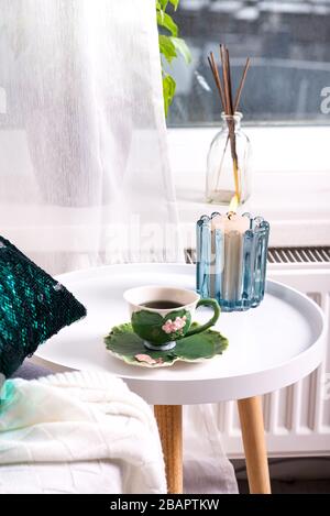 Eine Tasse Kräutertee auf einem Tisch, eine Strickwolldecke auf einem Stuhl und eine Kerze über dem Fenster. Schlafzimmer innen Stockfoto