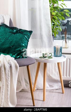 Eine Tasse Kräutertee auf einem Tisch, eine Strickwolldecke, ein grünes Paillettenkissen auf einem Stuhl und eine Kerze über dem Fenster. Schlafzimmer innen Stockfoto