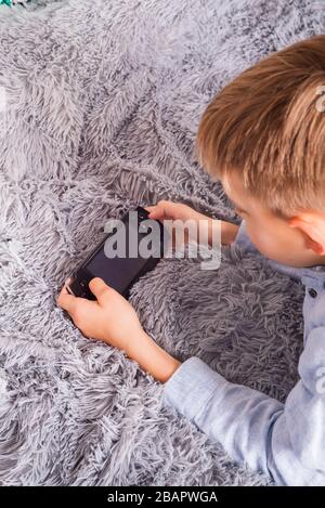 Kleiner Junge, der eine Handspielkonsole vor grauem Schaffelluntergrund spielt Stockfoto