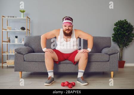 Lustiger fetter wütender Mann in der Sportswear sitzt auf dem Sofa im Zimmer Stockfoto