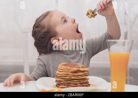 Süßes kleines Mädchen probiert Pfannkuchen mit weit geöffnetem Mund. Stockfoto