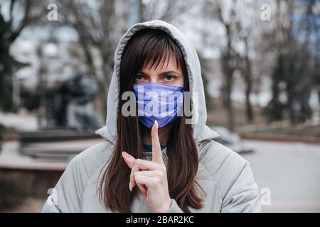 Frau mit einer medizinischen Maske im Gesicht hält den Zeigefinger am Mund und zeigt das Symbol leiser oder das oberste Geheimnis im Park Stockfoto