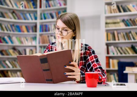 Attraktive blonde, intelligente junge Studentin in Brillen, die am Tisch sitzen, konzentriert beim Lesen des Buches in der modernen Universitätsbibliothek. Buchen Stockfoto