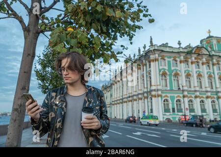 Die gutaussehenden Männer warten auf jemanden, trinken Kaffee und sehen im Telefon aus, er ist mit einer Militärjacke und Jeans bekleidet, das staatliche Hermitage-Museum ist eingeschaltet Stockfoto