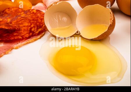 Zerbrochenes Hühnerei und Speck- und Wurstscheiben auf weißem Grund. Nahaufnahme Stockfoto