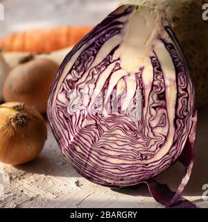 Ein halber Rotkohl auf einem rustikalen Tisch. Im Hintergrund Zwiebeln, Petersilienwurzel und Karotten. Gemüse für Salat oder Suppe. Vegane Ernährung. Gesunde Ernährung. Stockfoto