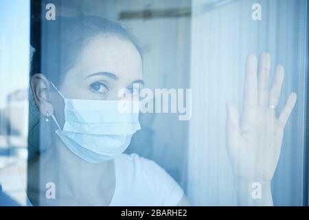 Das Mädchen in der medizinischen Maske ist im Krankenhaus unter Quarantäne gestellt und blickt durch das Glas in die Kamera. Blau getönt. Nahaufnahme. Stockfoto