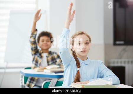 Portrait des süßen blonden Mädchens, das die Hand für die Antwort hebt, während es im Schulunterricht am Schreibtisch sitzt, Kopierer Stockfoto