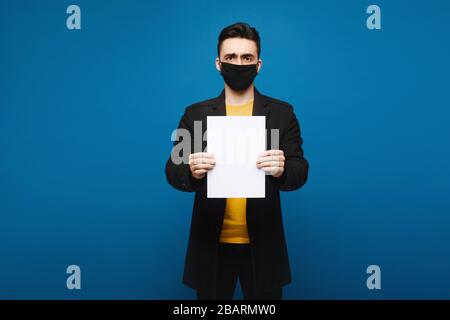 Junger Mann in einem schwarzen Mantel und schwarzer Schutzmaske, die mit einem leeren Blatt Papier am blauen Hintergrund posiert, isoliert mit Kopierraum. Werbeaktion Stockfoto
