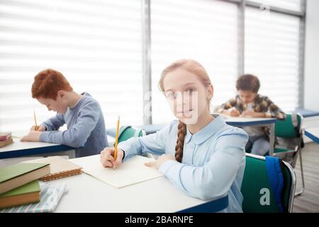 Portrait von süßen, intelligenten Schulmädchen, die im Klassenzimmer am Schreibtisch sitzen und die Kamera betrachten, während sie Notizen machen, Platz kopieren Stockfoto