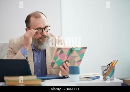 Portrait eines älteren Lehrers, das Buch liest, während er am Schreibtisch im Schulunterricht sitzt, Kopierer Stockfoto