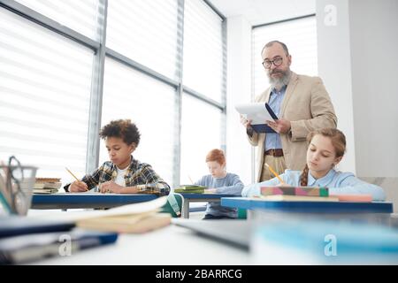 Portrait des reifen bärtigen Lehrers, der zwischen den Reihen läuft, während er Kindern beim Testen im Schulunterricht zuschaut, Kopierraum Stockfoto