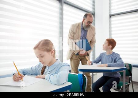 Gruppe von Kindern, die Test mit bärtigen, reifen Lehrern machen, die sie beobachten, konzentrieren sich auf süße blonde Mädchen, die am Schreibtisch sitzen und im Vordergrund schreiben, Kopie Spac Stockfoto