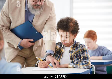 Portrait des bärtigen Oberlehrers, der afroamerikanischen Jungen hilft, im Schulunterricht am Schreibtisch zu sitzen Stockfoto